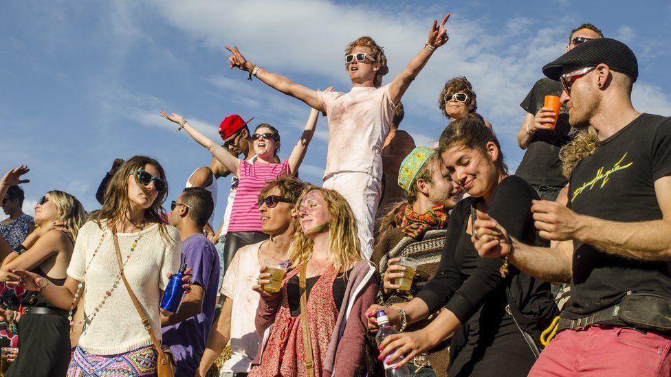 Multiple people raising their arms, wearing colourful outfits, singing along to songs at Secret Garden Festival. People are holding drinks, water bottles, wearing sunglasses and hats, while the sun is shining and the sky is blue with a shade of cloud.