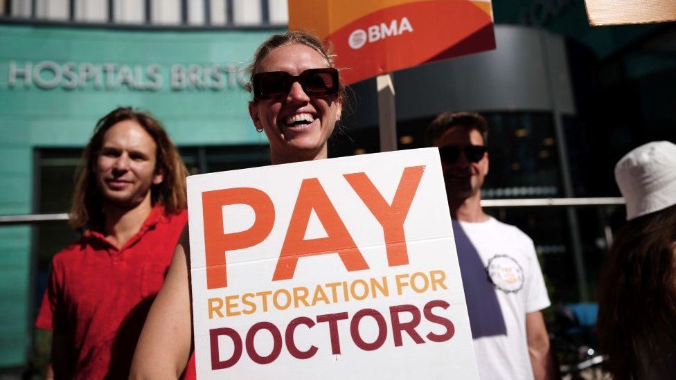 Photo of a striking doctor in glasses holding a placard, which reads 'pay restoration for doctors'