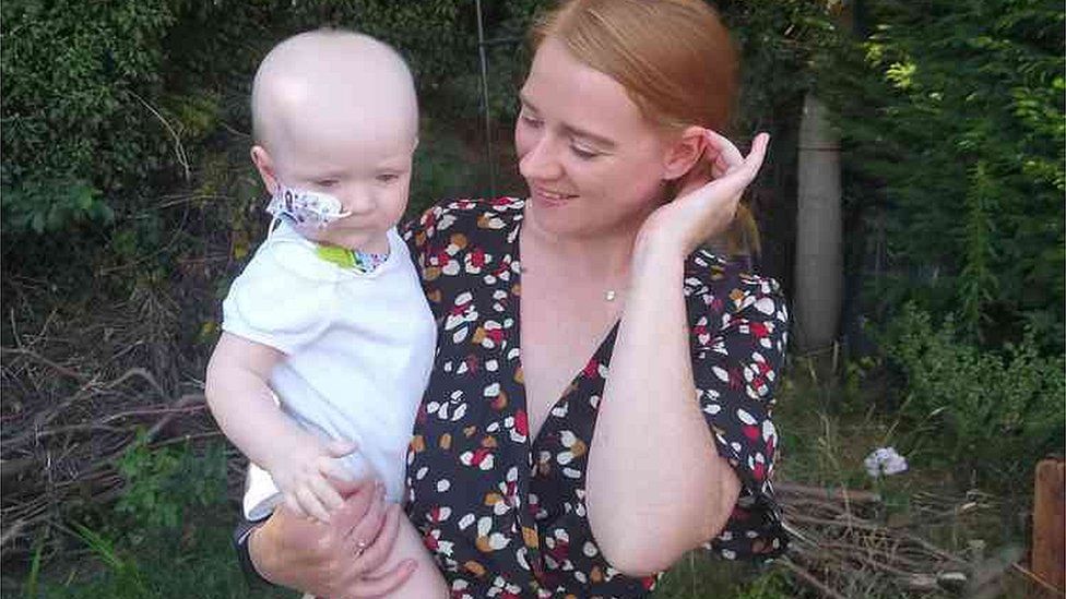 Callan Brett, 33, of Ipswich, with her baby son Henry in a woodland setting. Baby Henry has a breathing tube affixed to his nose and wars a white Babygro. Callan is stroking her hair behind her ear and is smiling at her son